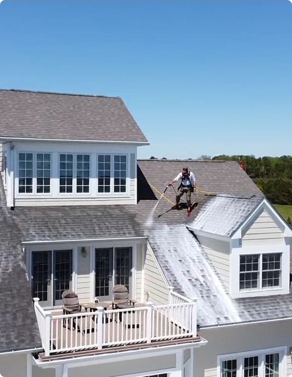 A person wearing a harness is cleaning the roof of a large house with a long pole, spraying a white substance, on a sunny Florida day. Below, the balcony has chairs and a table—just another day for Sunny Side Roof Rejuvenate roofing experts.