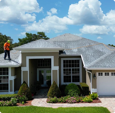 A worker wearing safety gear stands on the roof of a suburban Florida house, using equipment to clean or inspect the roof under a bright, partly cloudy sky. Sunny Side Roof Rejuvenate ensures roofing stays pristine amid well-maintained landscaping.